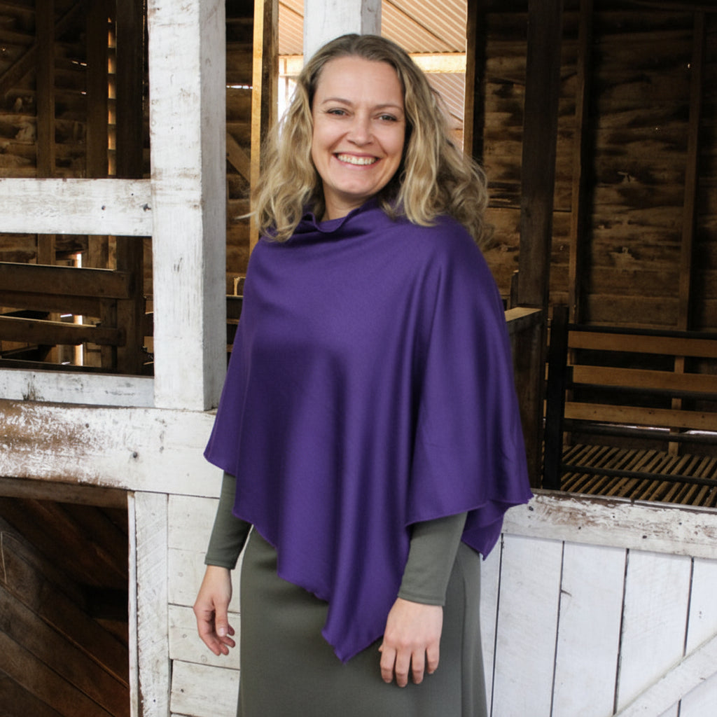 A smiling woman with blonde hair models a draped purple shawl or poncho over her shoulders, worn over an olive green long-sleeve top and skirt, set against a rustic wooden barn interior.