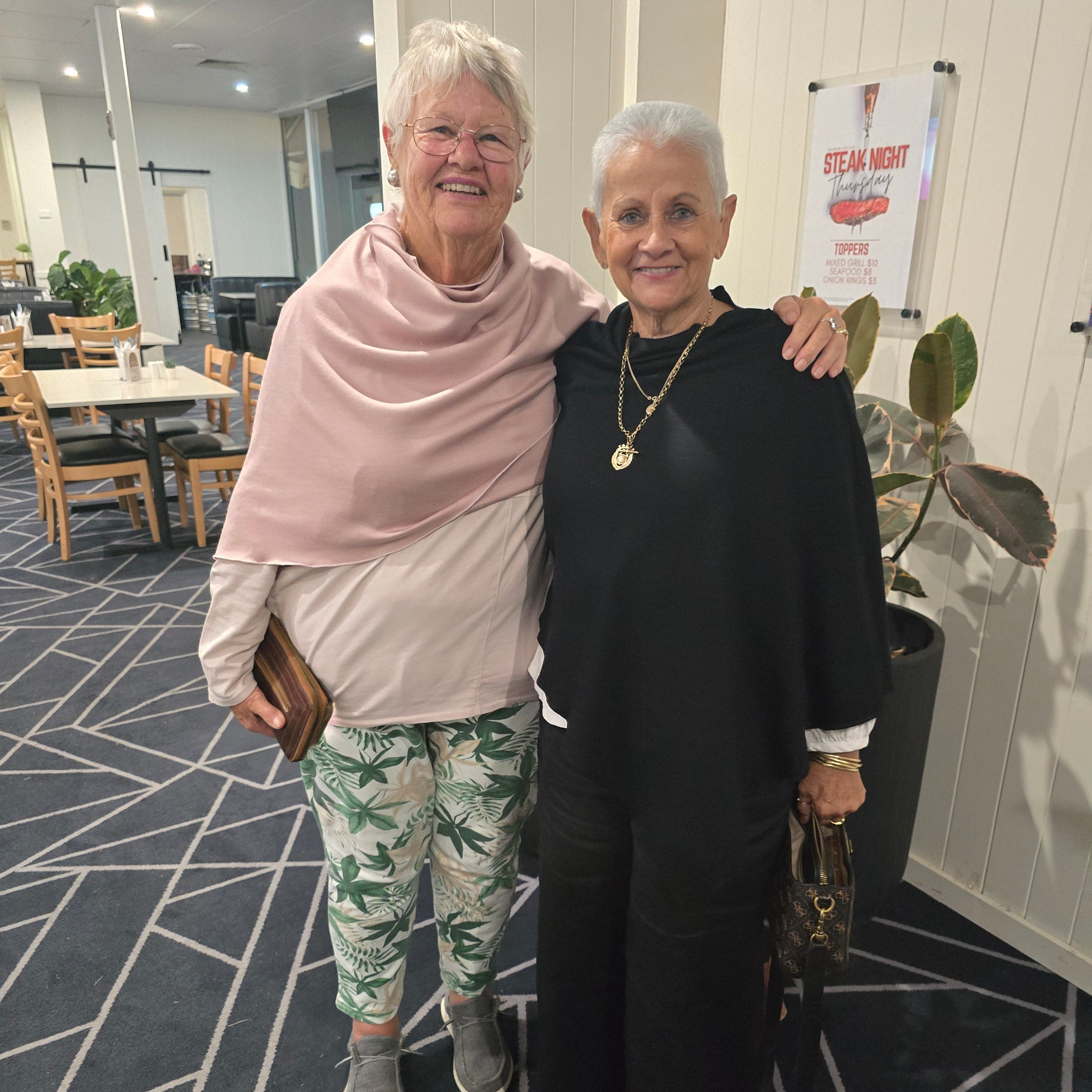 Two elderly women posing together indoors. The woman in the foreground wears a light pink top and green palm-patterned pants, while the woman behind her wears a black long-sleeve top and black trousers.
