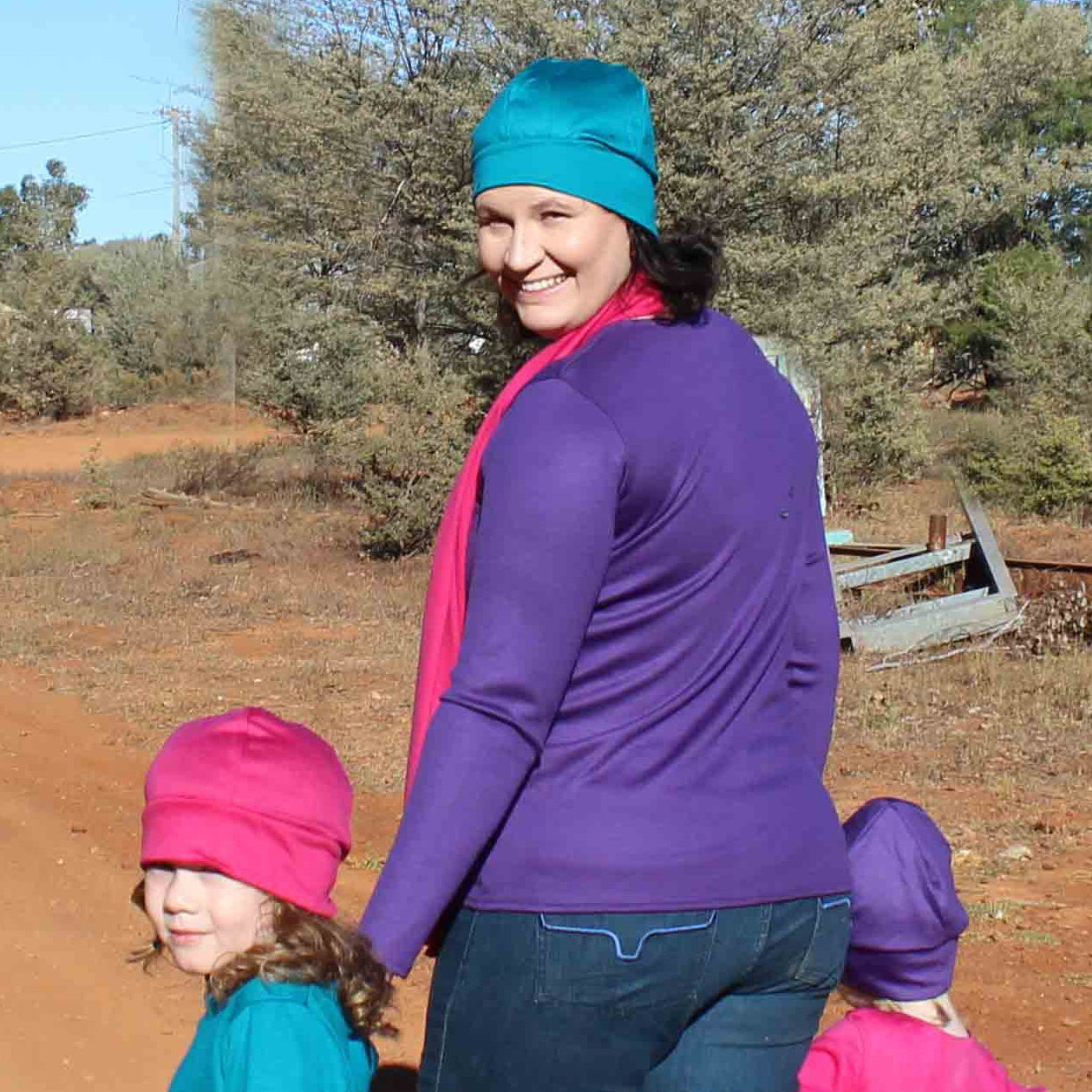 Woman in a purple jacket and blue beanie with two children outdoors on a dirt path.